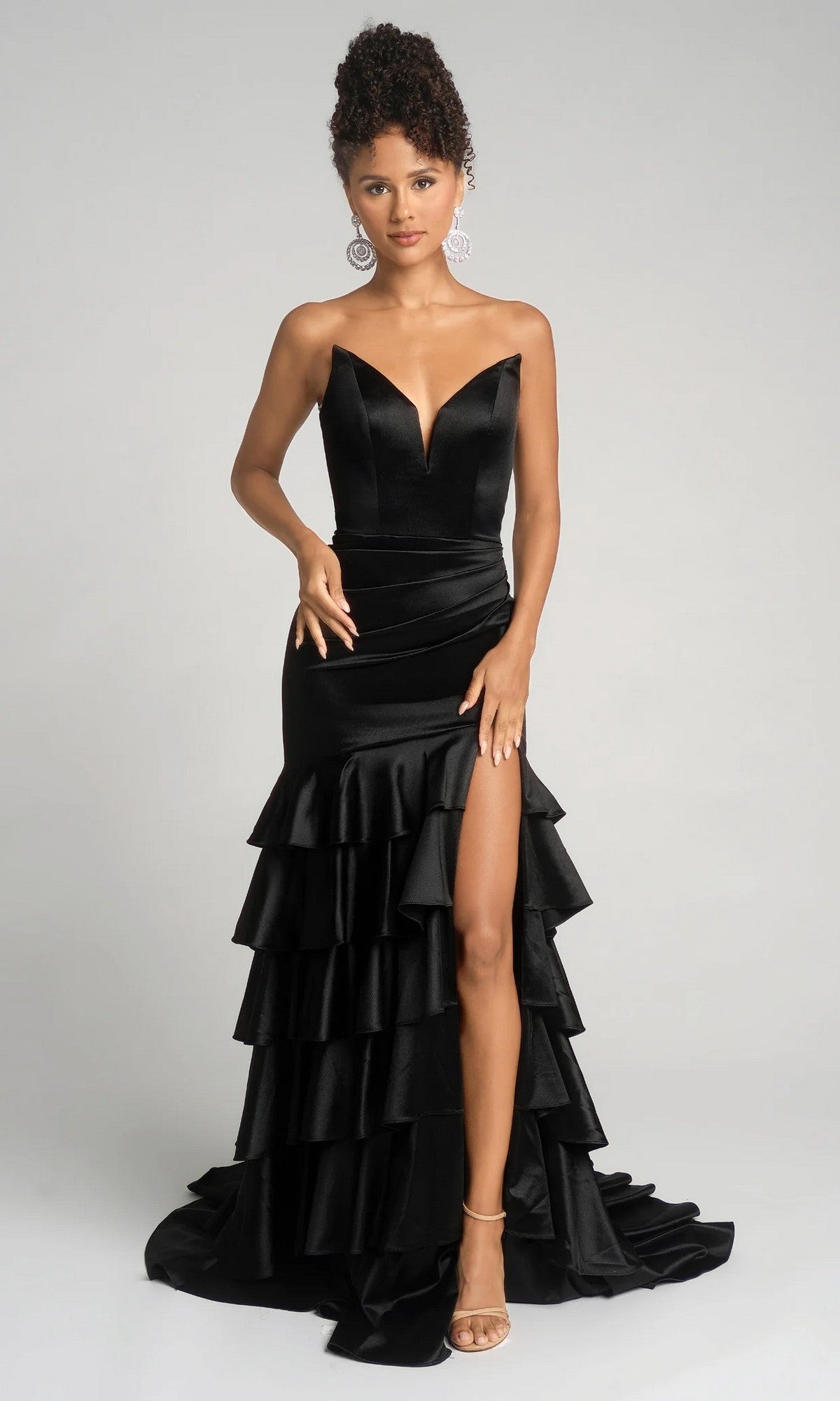 A woman poses against a plain background in the Portia and Scarlett XO021 long prom dress—a black gown with a strapless deep V-neckline, tiered ruffle skirt, and high slit.